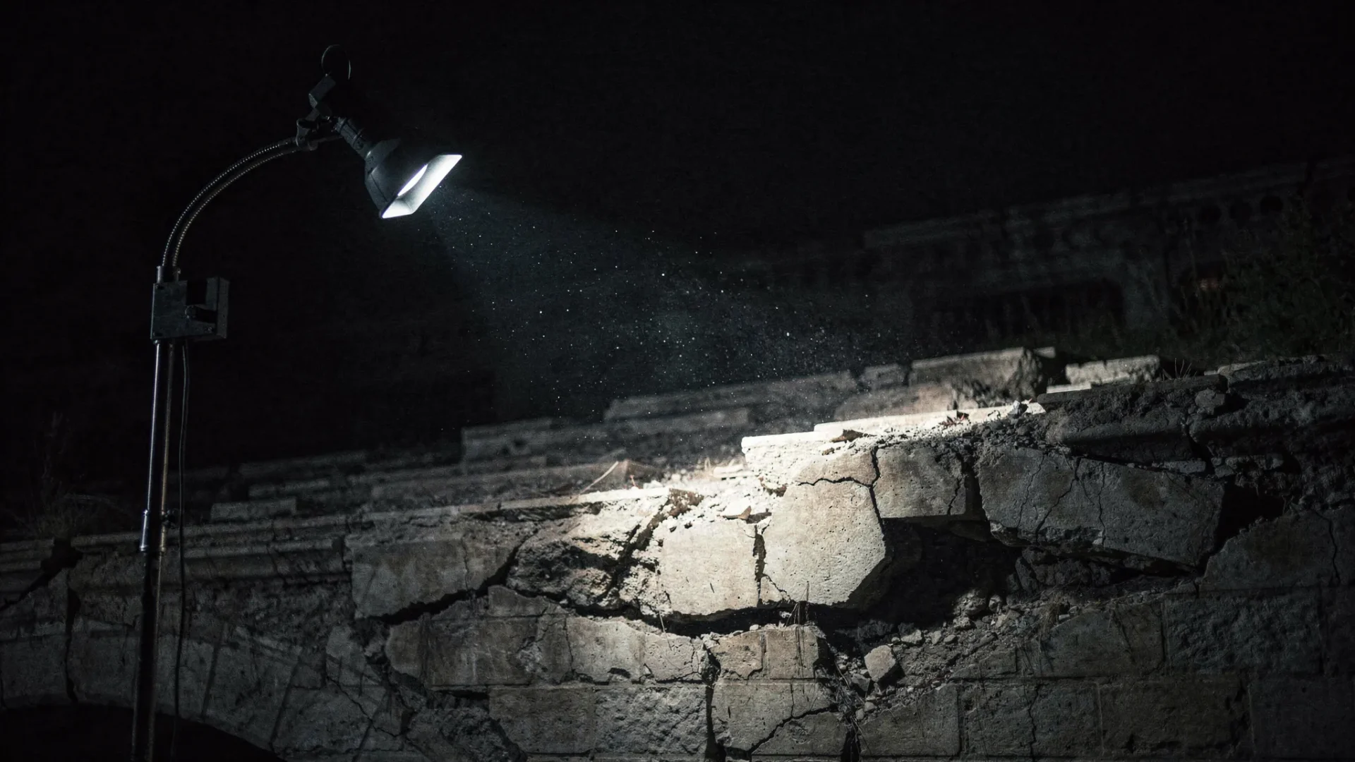 A single inspection lamp cuts through darkness across a cracked stone bridge, revealing hidden faults beneath the surface while the surrounding space fades into shadow. The scene feels tense, quiet and forensic, with deep contrast and a strong focal point on the damaged structure.