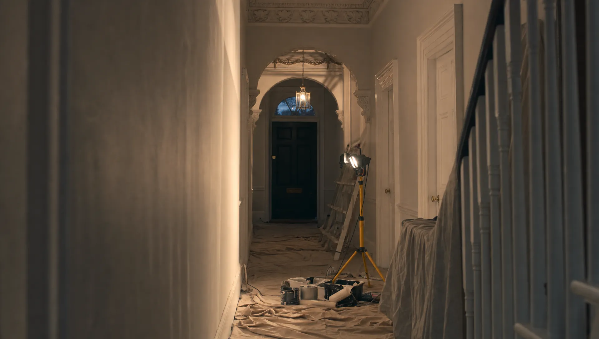 A freshly painted Georgian hallway at dusk, with a single work light casting dramatic shadows across dust sheets, paint tins and crisp white woodwork, showing careful preparation and tidy local workmanship.