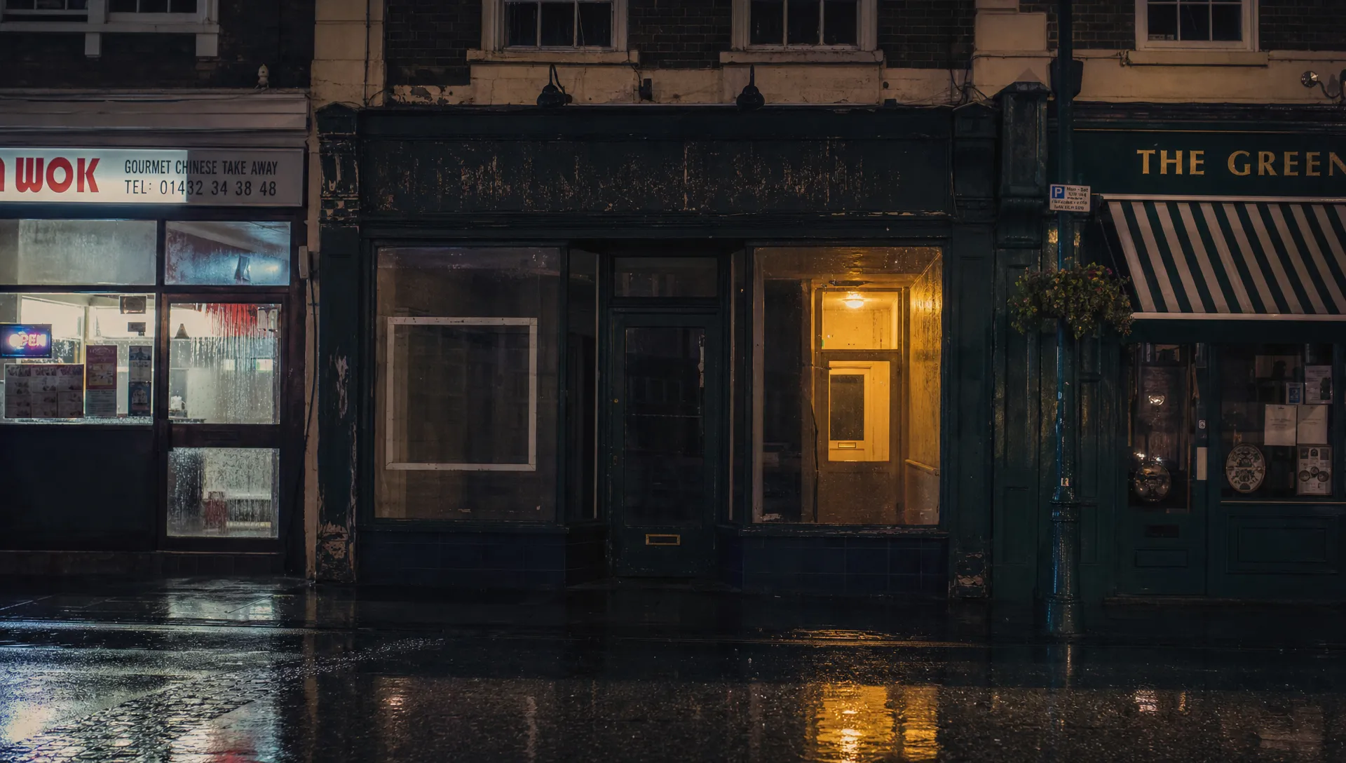 An empty shopfront on a quiet British high street at night, with one warm light glowing from a back room while brighter neighbouring signs reflect in rain on the pavement.