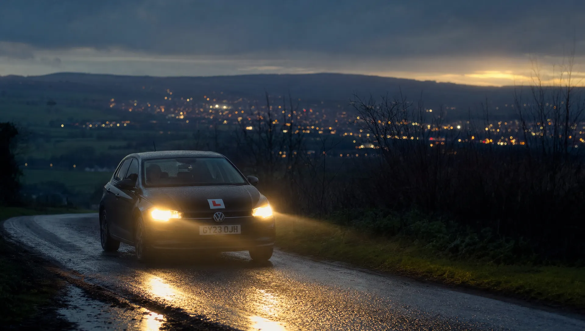 A learner car on a wet country road at dusk, with headlights cutting through mist and town lights glowing ahead.