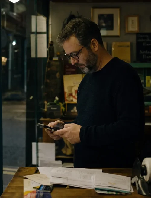 A shop owner checks business details on a phone beside invoices and receipts after closing time.