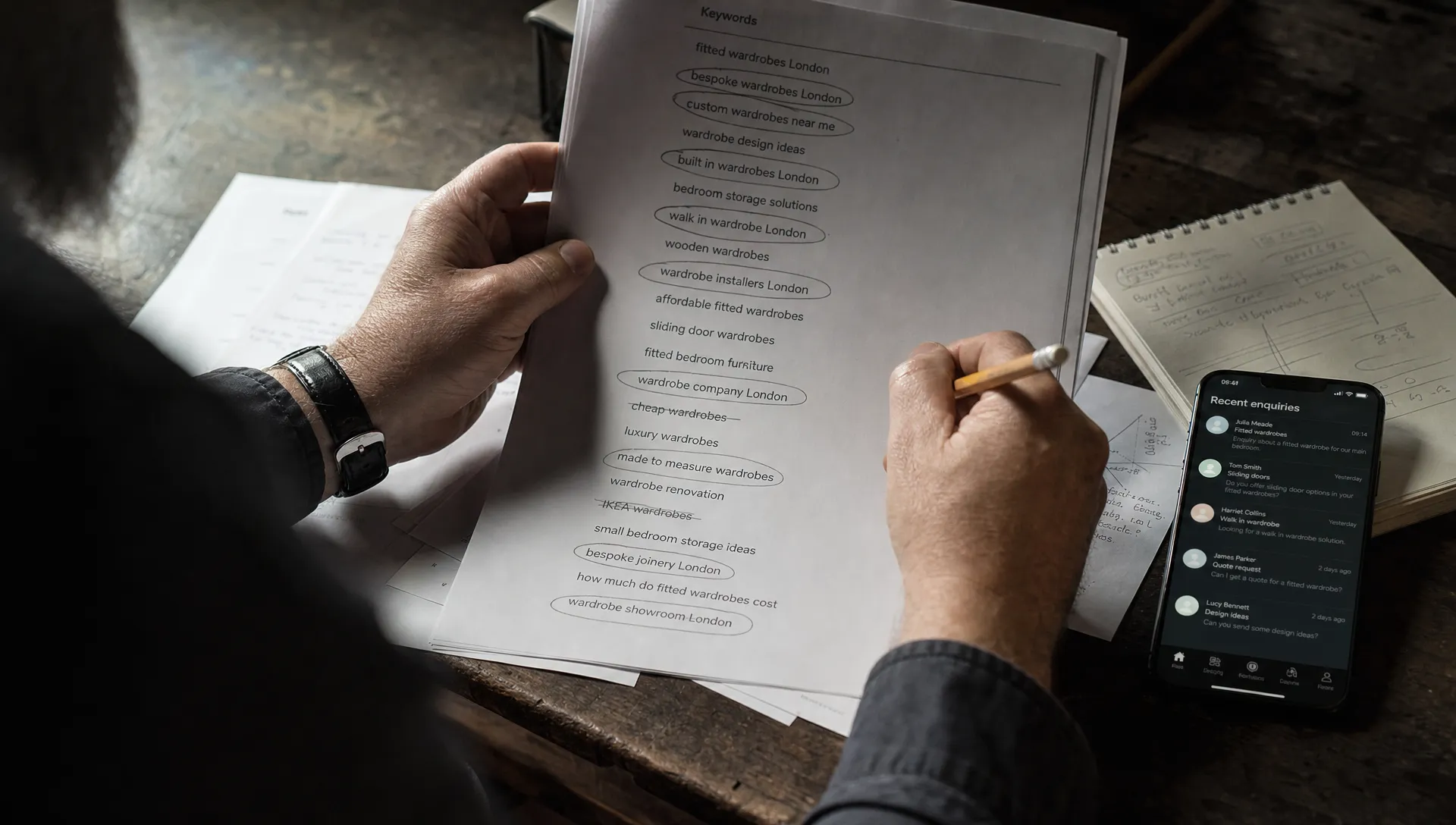Hands sort printed search phrases beside a phone and notebook, showing the work of choosing useful long-tail keywords.